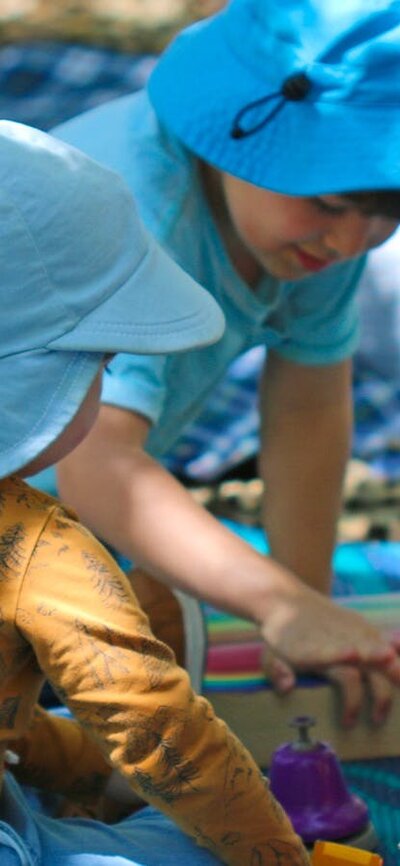 Three preschool children sit on picnic mats on the forest floor playing with wooden blocks