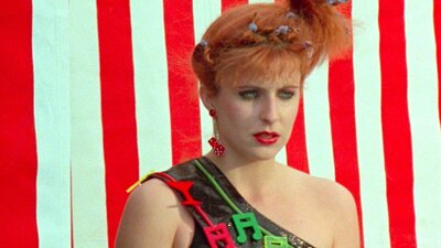 NFSA Restores: Starstruck (1981) woman with red lipstick and 80's hairstyle looks glum past the camera, red and white stripe backgrou