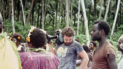 One cameraman stands looking in his pocket, other actors standing around