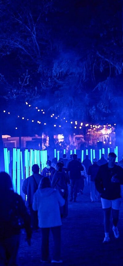 Visitors walk along a path lined with glowing blue light poles at a night time festival.