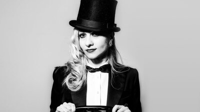 Nikki Nouveau in top hat, bow tie and tails, straddles a chair, facing the viewer.