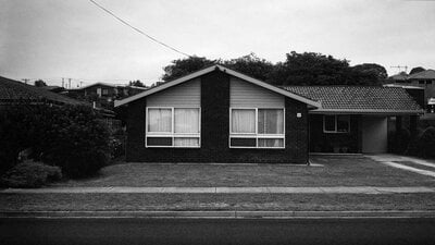 A black and white photograph of a suburban scene featuring a house, a bare front yard and street