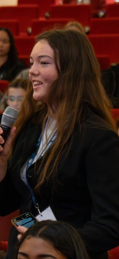 Photo in theatre, in focus is a conference attendee with a microphone asking a question