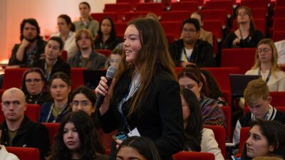 Photo in theatre, in focus is a conference attendee with a microphone asking a question
