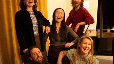 5 performers laughing in front of a black piano and golden theatre curtains