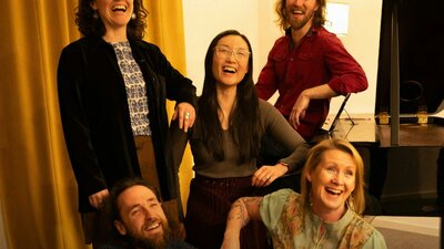 5 performers gathered together laughing next to a grand piano