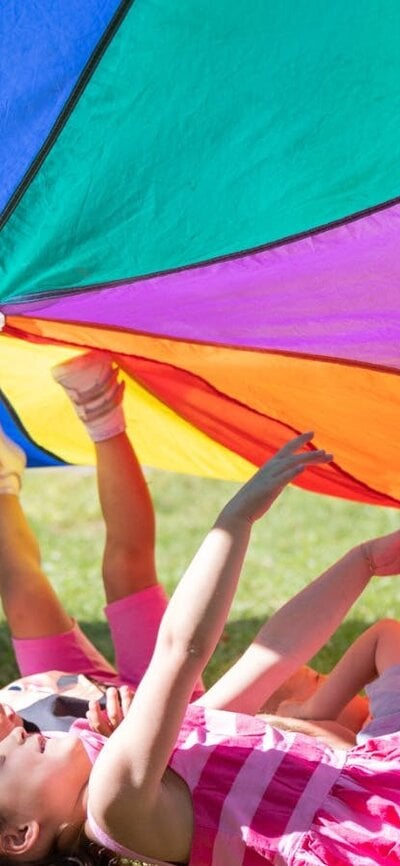 A group of children laying in the grass under a rainbow parachute.