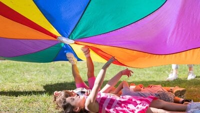 A group of children laying in the grass under a rainbow parachute.