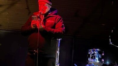 Man on stage wearing a beanie, holding a mobile phone and speaking into a microphone