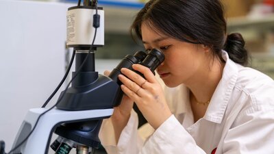 Learn how to operate a microscope and view parasite samples prepared by scientists at ANU! A girl in a white lab coat looks into the eyepiece of a microscope.