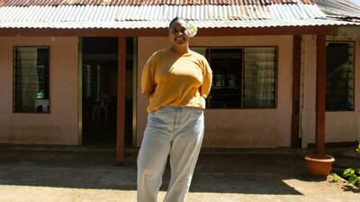 A smiling woman in a yellow shirt and light blue pants stands in front of a house.