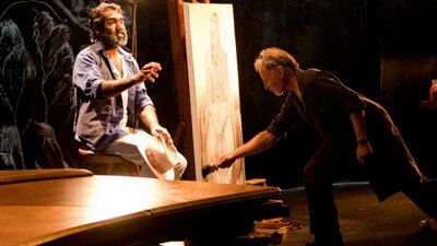 Two people on a darkened stage, one seated beside an easel while the other paints a portrait.