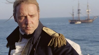 Man dressed as a captain stands with wind through his hair and sun on his face, ship & water behind