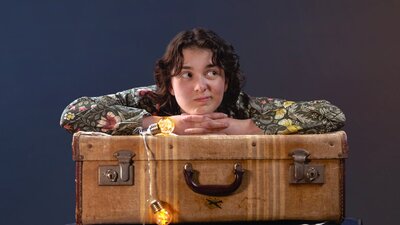 Peter/Wendy Banner A young woman with short dark curls rests her chin on her hands on top of a brown vintage suitcase.