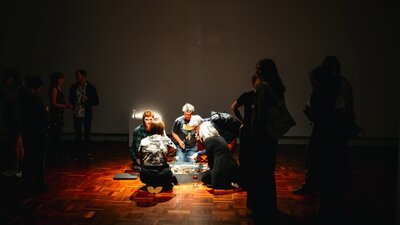 Visitors gather around a spotlit, art installation in a dimly lit exhibition space.