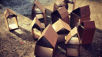A cluster of small cardboard houses, in sunlight on a gravel surface.