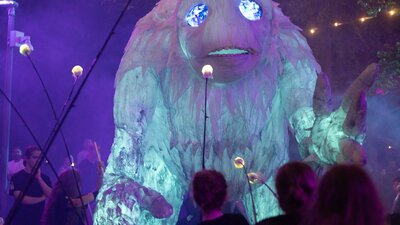 A large, mysterious Yeti puppet strides through a crowd of people at night.