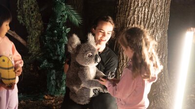 A child interacts with a koala puppet, manipulated by a puppeteer, in a forest at night.