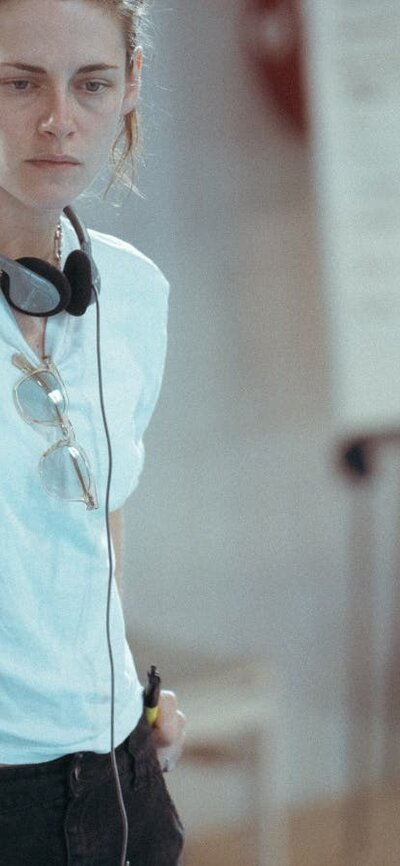Woman stands with headphones and white t shirt with hands in pockets looking forlorn