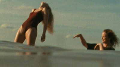 Woman in bathing suit standing in water leaning back with another woman flicking water at her