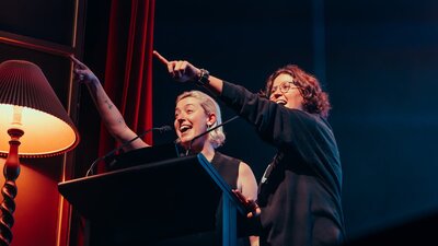 Hosts Harriet Gillies and Xanthe Dobbie on-stage behind a lectern, joyously pointing at audience.