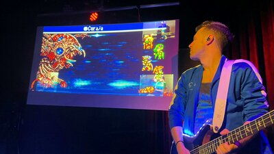 A man playing a guitar standing next to a TV screen with a video game