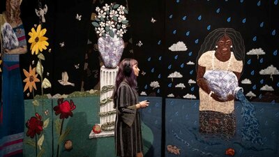 Tal Fitzpatrick | Quietly Seething: Fate, Fury and Transmutation A woman stands in front of a large tapestry artworks with flowers and a woman with a water vessel