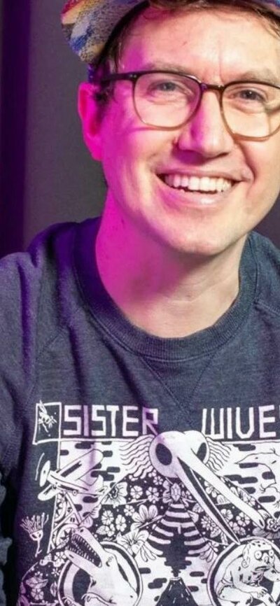 Fionn McCabe A smiling man wearing glasses and a colorful cap, seated in front of a dark curtain.