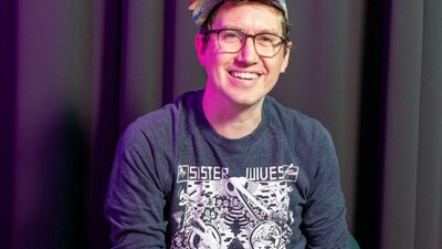 Fionn McCabe A smiling man wearing glasses and a colorful cap, seated in front of a dark curtain.