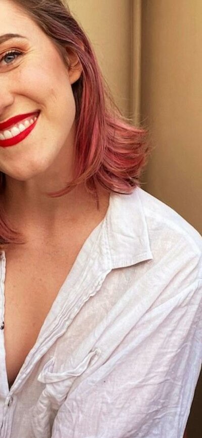 Evie Hilliar A smiling woman with brown hair with pink highlights sits on a wooden bench outdoors.