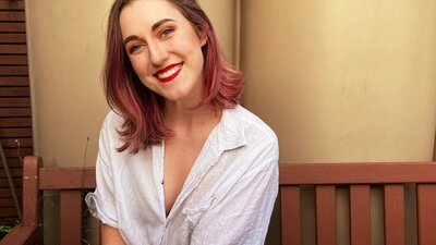 Evie Hilliar A smiling woman with brown hair with pink highlights sits on a wooden bench outdoors.