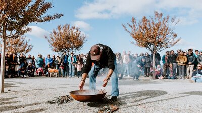 Person tending to a fire pit during a smoking ceremony, surrounded by a large crowd