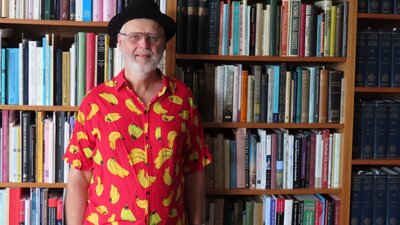 An elderly man wearing a black hat and a banana printed shirt while standing in front of a bookcase