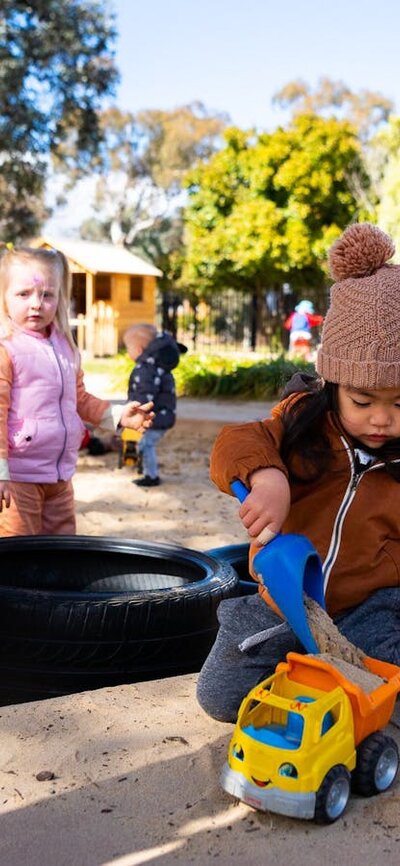 Young children play in a sandpit and look at the camera.