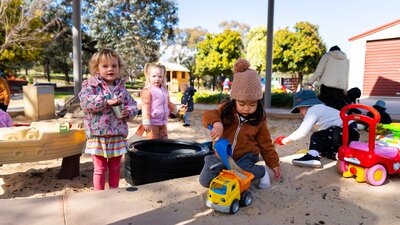 Young children play in a sandpit and look at the camera.