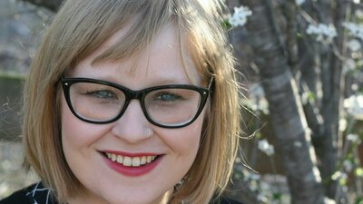 A smiling woman with glasses and blonde hair stands outdoors near blooming trees.
