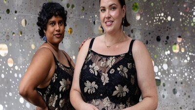 Two women posing confidently in black and gold lace dresses, celebrating diverse bodies.