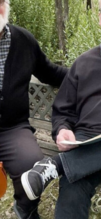 Two elderly men sitting on a bench in nature, one holding a viola