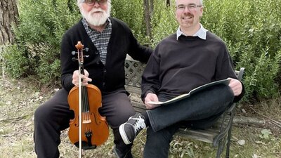 Two elderly men sitting on a bench in nature, one holding a viola