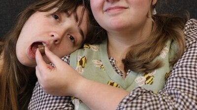 Two young women sit next to each other. One of the women feeds the other a chocolate.