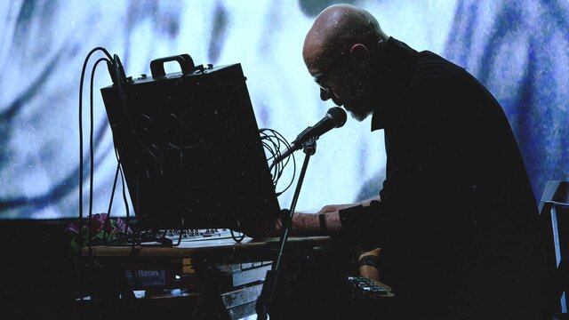 Performer sits at audio equipment, speaking into a microphone before a glowing blue projection.