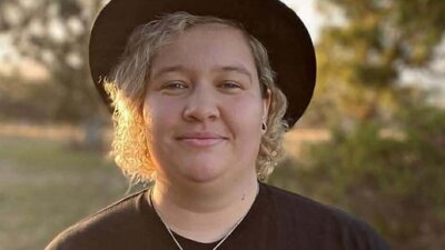 A person with curly blonde hair wearing a black hat and black t-shirt smiles at the camera.