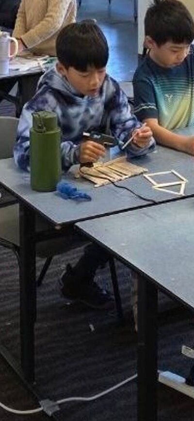 Students working on their projects A photo of students using glue and craft sticks to build contraptions, with a miscroscope on a table