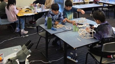 Students working on their projects A photo of students using glue and craft sticks to build contraptions, with a miscroscope on a table