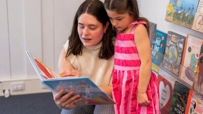 A woman reading a book to a girl in a pink dress