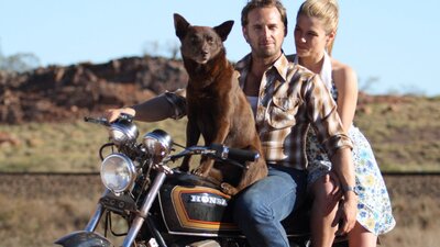 Still from Red Dog showing a man, a woman and a kelpie on a motorcycle in an outback landscape