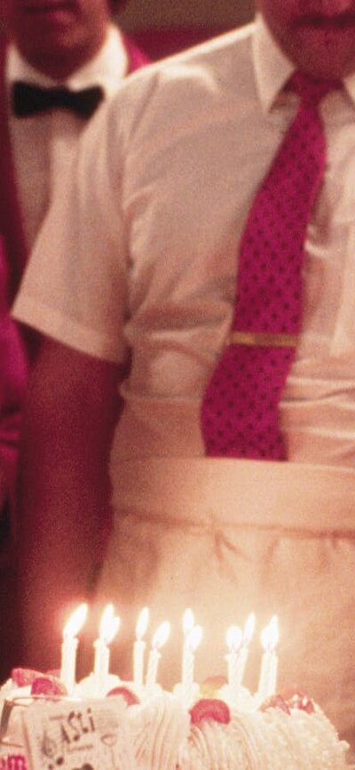 Man and boy sit at a table with a cake with candles in front and wait staff standing