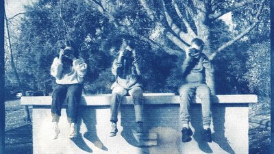A cyanotype print photograph of three kids sitting on a ledge holding cameras up to their faces