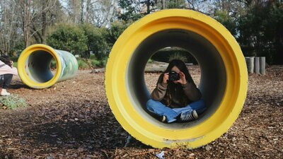 Person sitting cross-legged inside a yellow concrete pipe in a park, aiming a camera at the viewer.