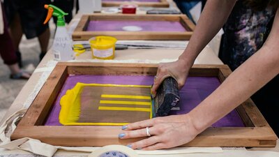 Hands rolling yellow ink onto a purple screen-printing frame with a squeegee at a workshop table.